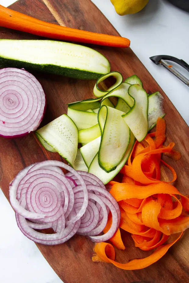 Shaved carrots and zucchini with thinly sliced red onion on a cutting board. 