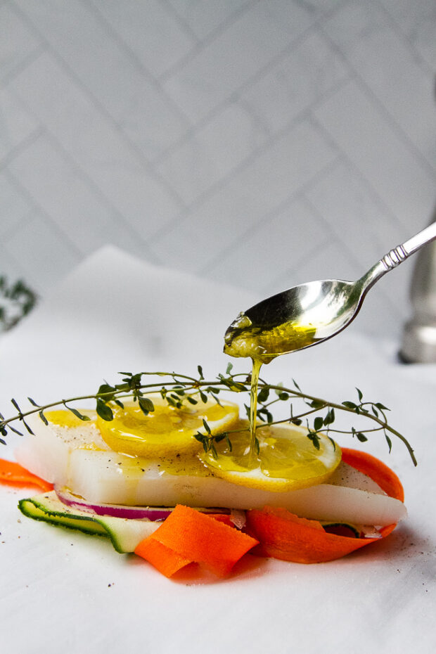 Layers of thinly sliced vegetables, fish, citrus, and herbs with a drizzle of garlic olive oil on parchment. 