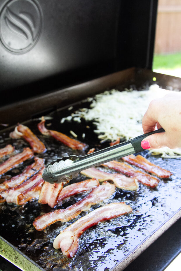 Flipping bacon with a pair of tongs on a Blackstone griddle.