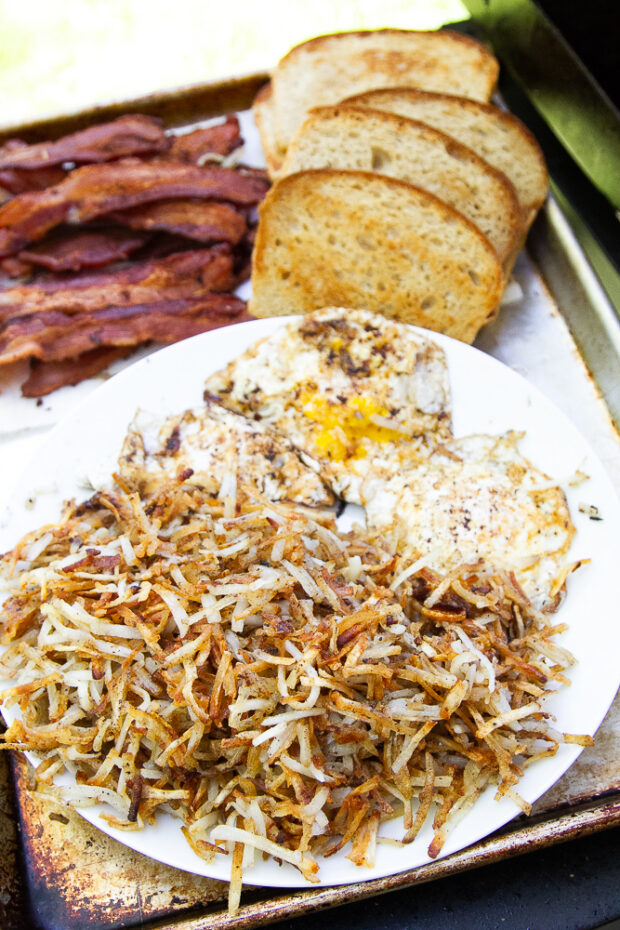 A platter with cooked bacon, hash browns, fried eggs, and toast.