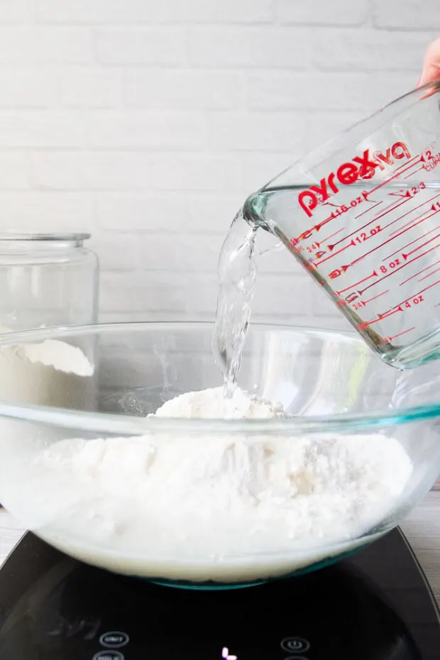 Glass bowl on top of a kitchen scale with flour inside. Water is being poured into the bowl. 