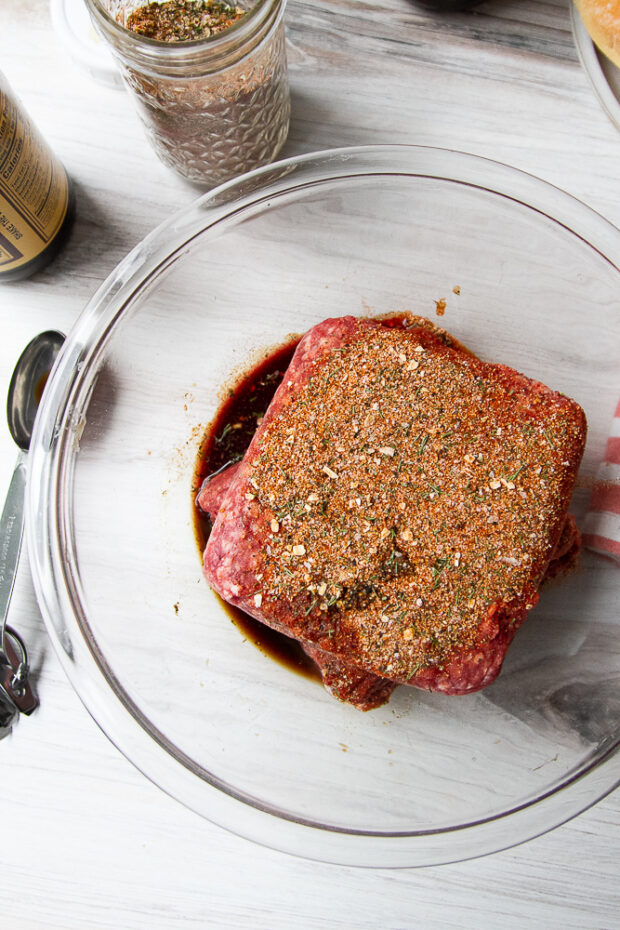 Ground beef in a clear bowl with steak seasoning and Worcestershire sauce added.