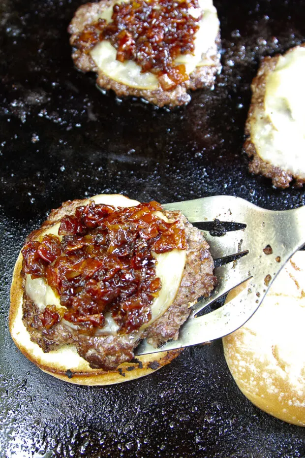 Cooked bacon patties on a griddle with melted cheese and bacon jam. A patty is being placed on a toasted bun.