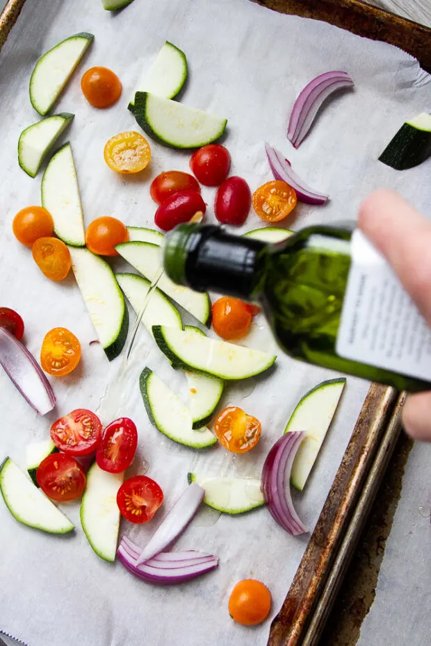 A parchment lined sheet pan with chopped zucchini, tomatoes, and red onion. Avocado oil is drizzled over the top.