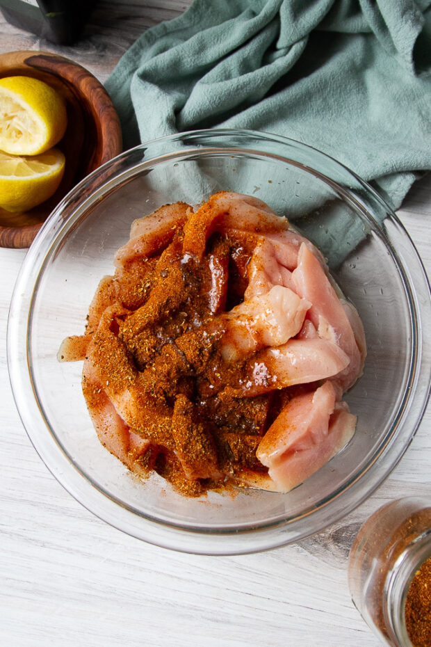 Clear bowl with uncooked sliced chicken with shawarma seasoning, lemon juice, and oil added on top.