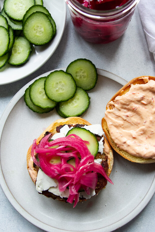 A plate with an open burger filled with toppings and some cucumber slices on the side. 