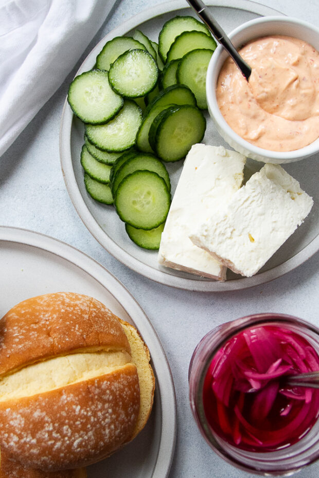 A plate with chopped cucumbers, feta cheese, and a small bowl of spicy mayo. Alongside sits the jar of pickled red onions, and a plate with buns. 