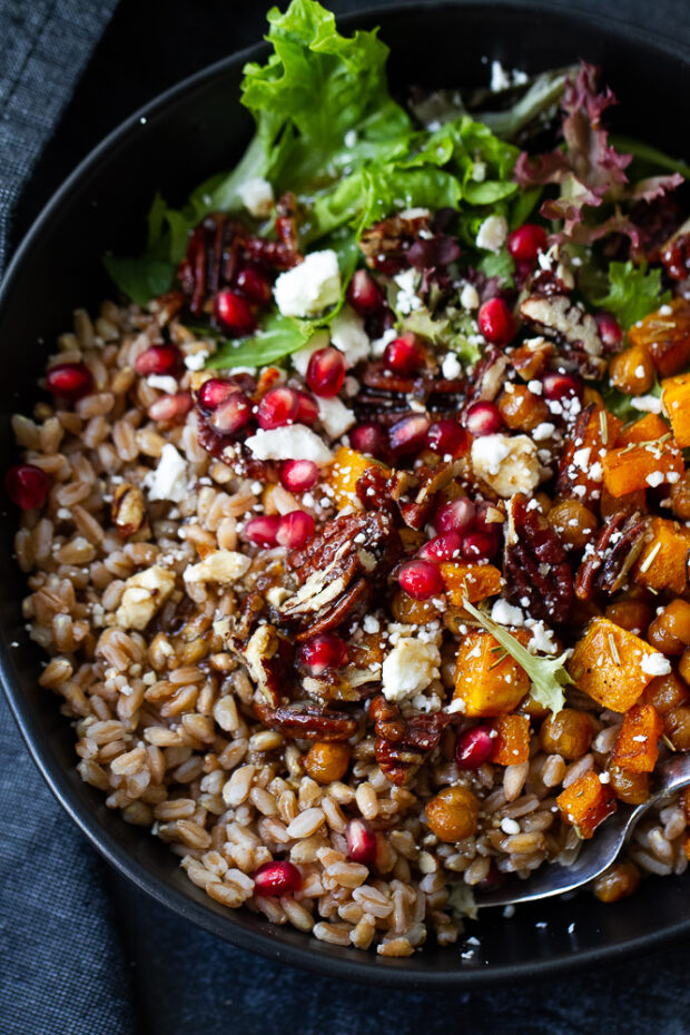 Buddha bowl in a black bowl with farro, greens, roasted butternut squash, chickpeas, feta, pomegranate arils, pecans, and a balsamic vinaigrette.