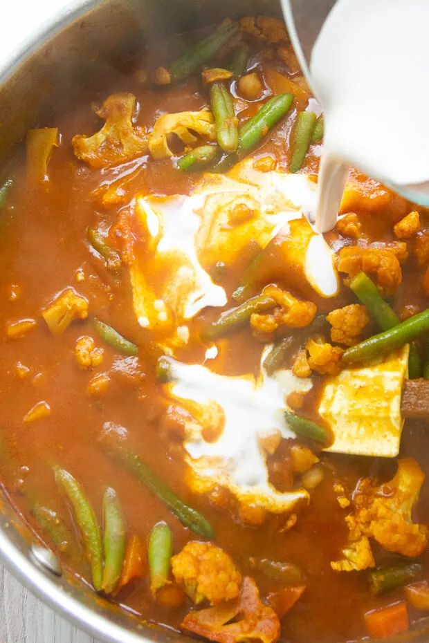 Curry has simmered and thickened and coconut milk is being added to the skillet.