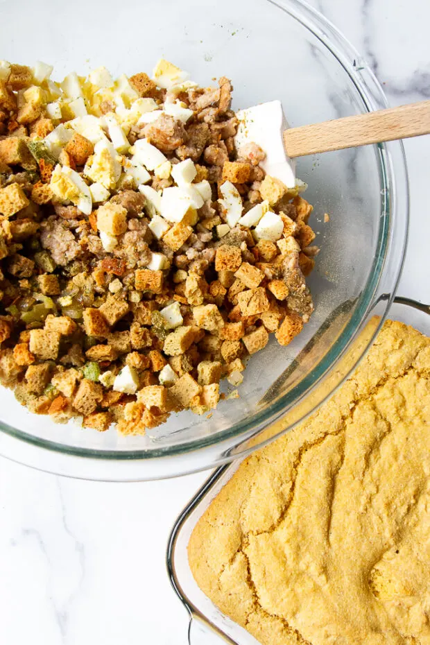 Baked cornbread next to a clear bowl with the rest of the dressing ingredients (cooked sausage, bread cubes, onions, celery, hardcooked eggs)